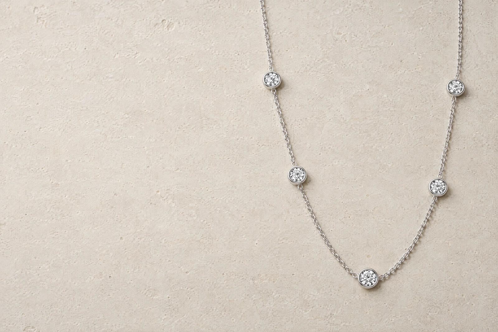 The Classic Station Necklace laid in a deep U on pale limestone, its five round clear stones spaced along a fine silver chain