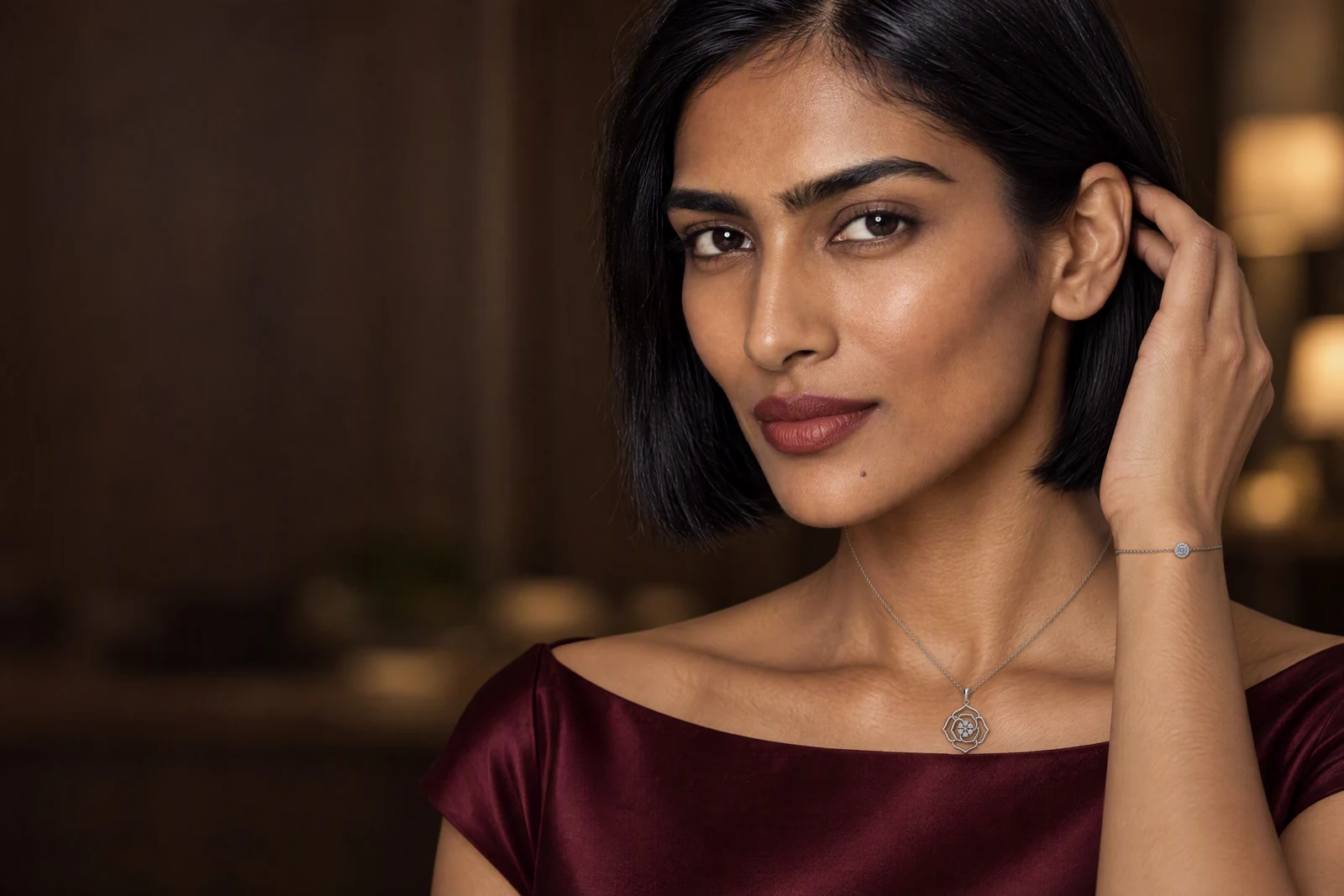 A woman with a short bob in a wine satin top in a lamplit room, her hand raised to her hair, wearing the Bloom Multi Stone Pendant and the Celeste Halo Bracelet