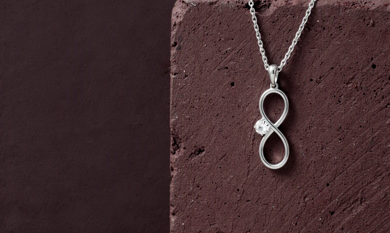 The Infinity Solitaire Pendant on its chain, hanging over the edge of a plum plaster block