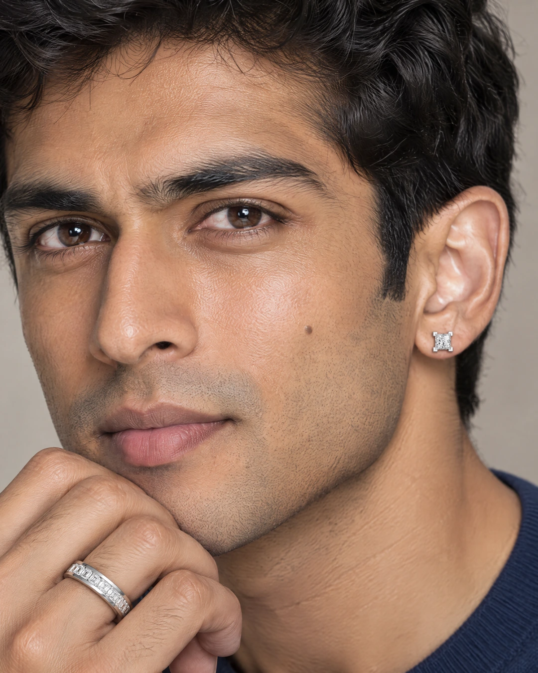 A man in a navy sweater on a pale stone backdrop, his jaw resting on his hand, wearing the Square Eternity Ring and a princess-cut Square Stud Earring