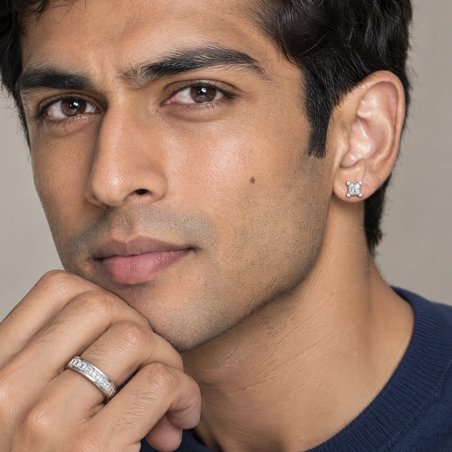 A man in a navy sweater, his jaw resting on his hand, wearing the Square Eternity Ring and a Square Stud Earring