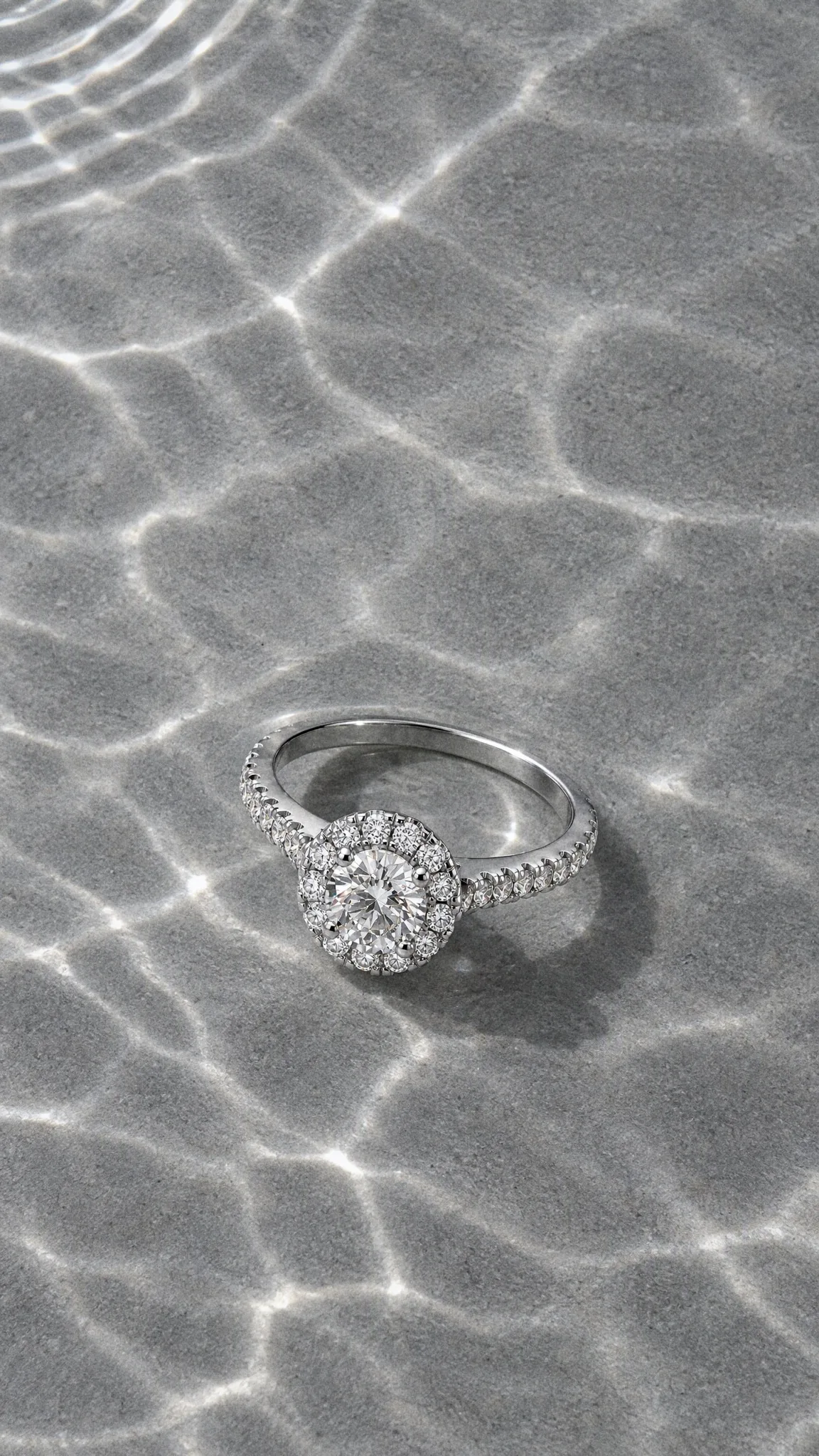The Celeste Halo Ring lying in shallow water over grey sand, light rippling across it