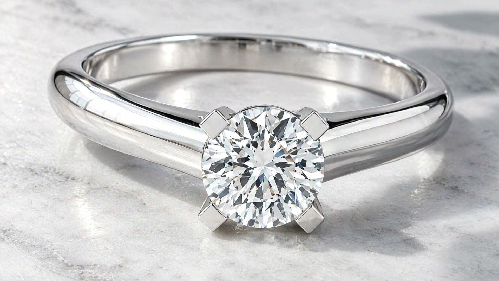The Classic Solitaire Ring on white marble in morning sun, close on its one round stone held in four claws