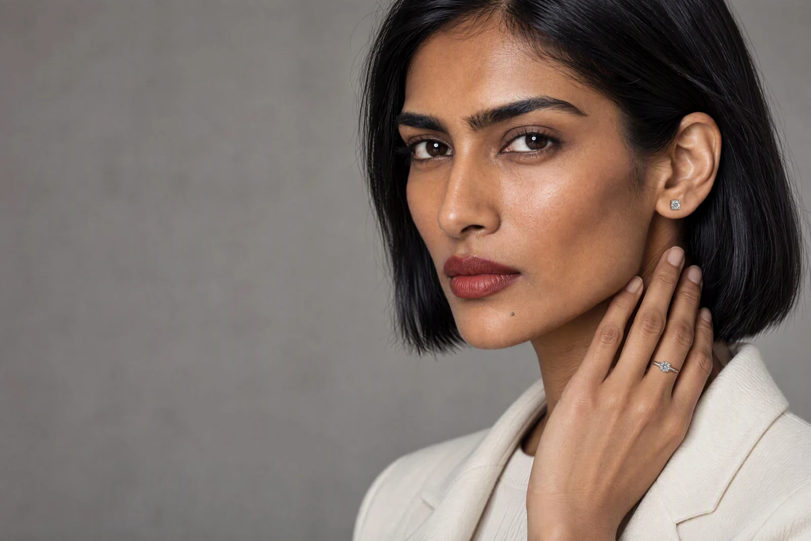A woman with a short bob in an ivory blazer against a grey wall, her hand at her jaw, wearing the Classic Solitaire Ring and the Classic Stud Earrings