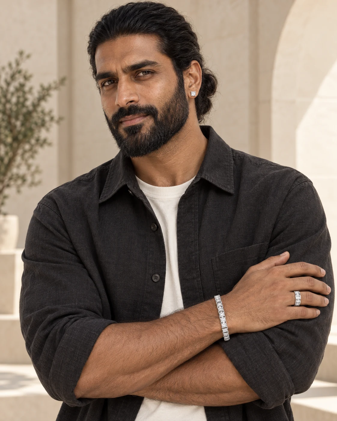 A bearded man with tied-back hair and relaxed folded arms. An Indian man wearing the Square Eternity Ring, princess-cut Square Stud Earrings and the Emerald-Cut Tennis Bracelet together